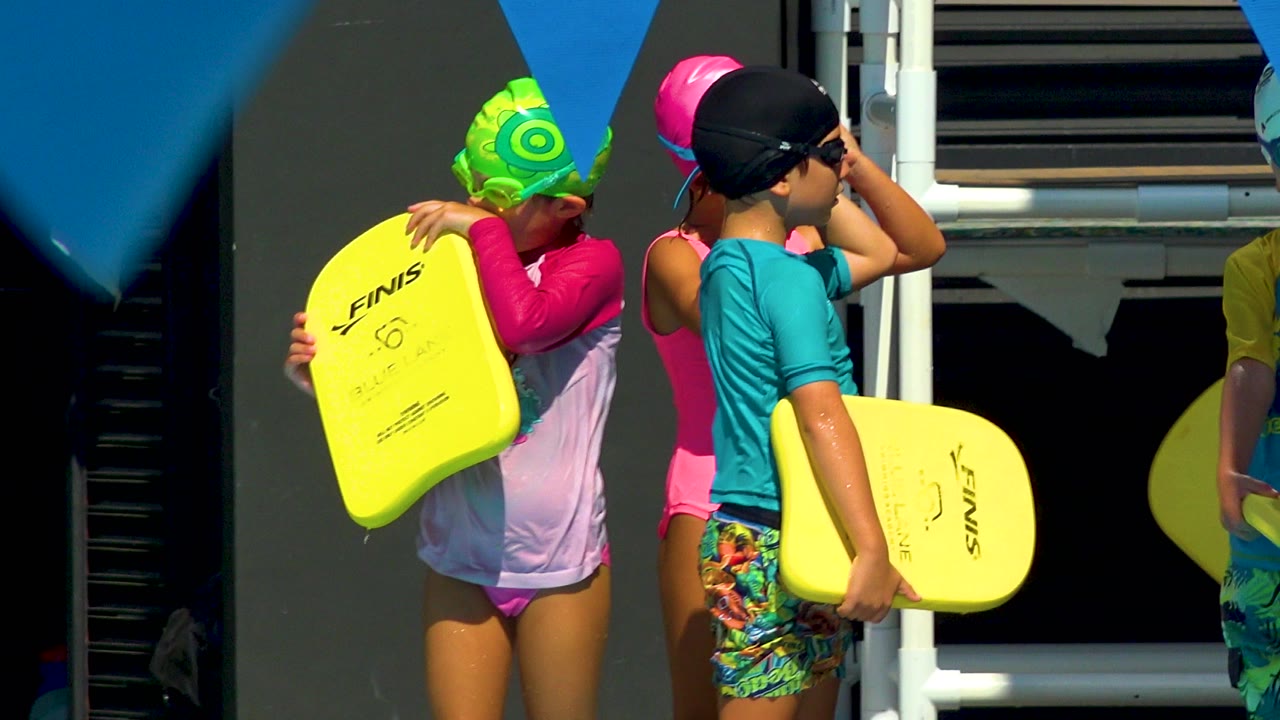 Children adjust goggles while holding yellow swim kickboards.
