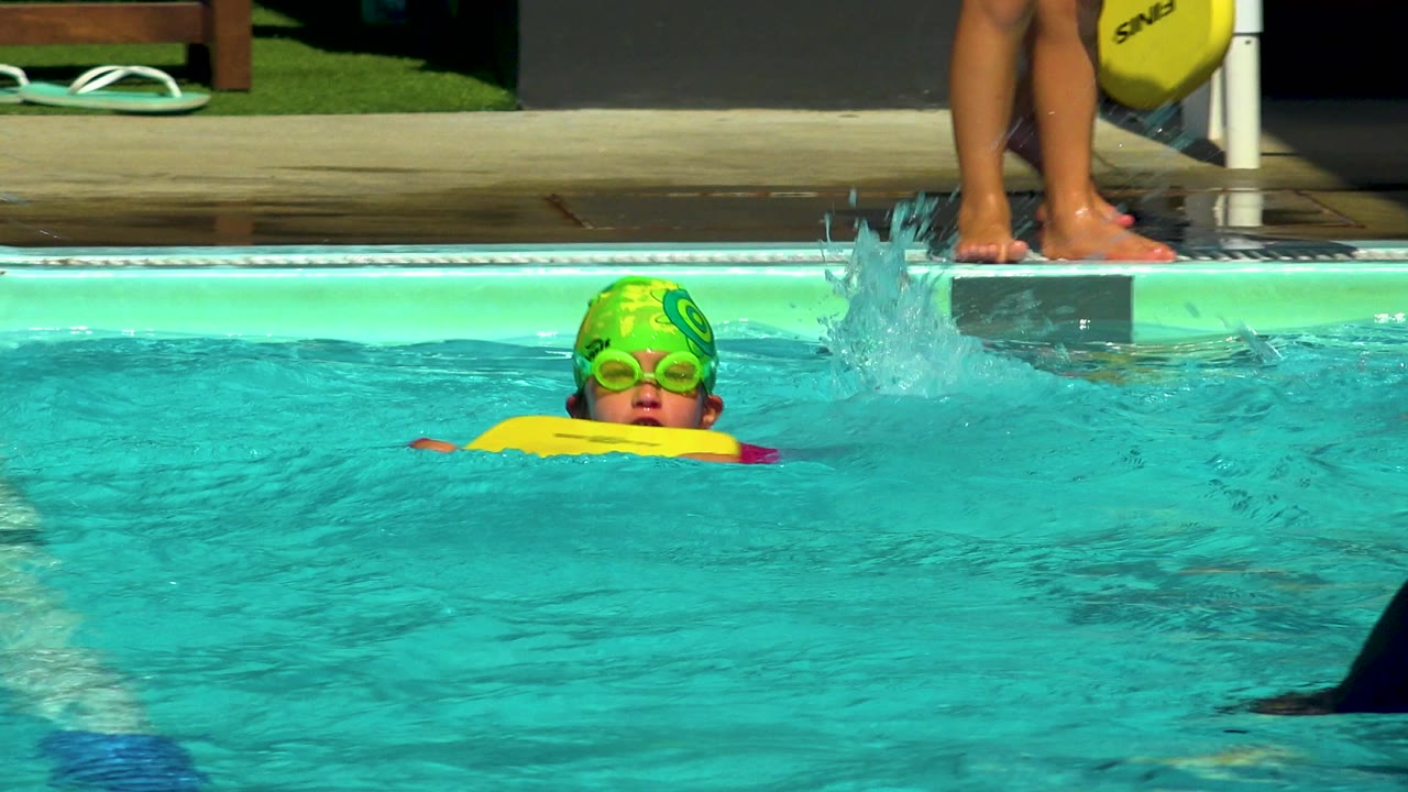 At 0:00, a child in a green swim cap and goggles faces camera while holding a yellow kickboard in a Cabo San Lucas pool, with an adult's feet visible on the deck.