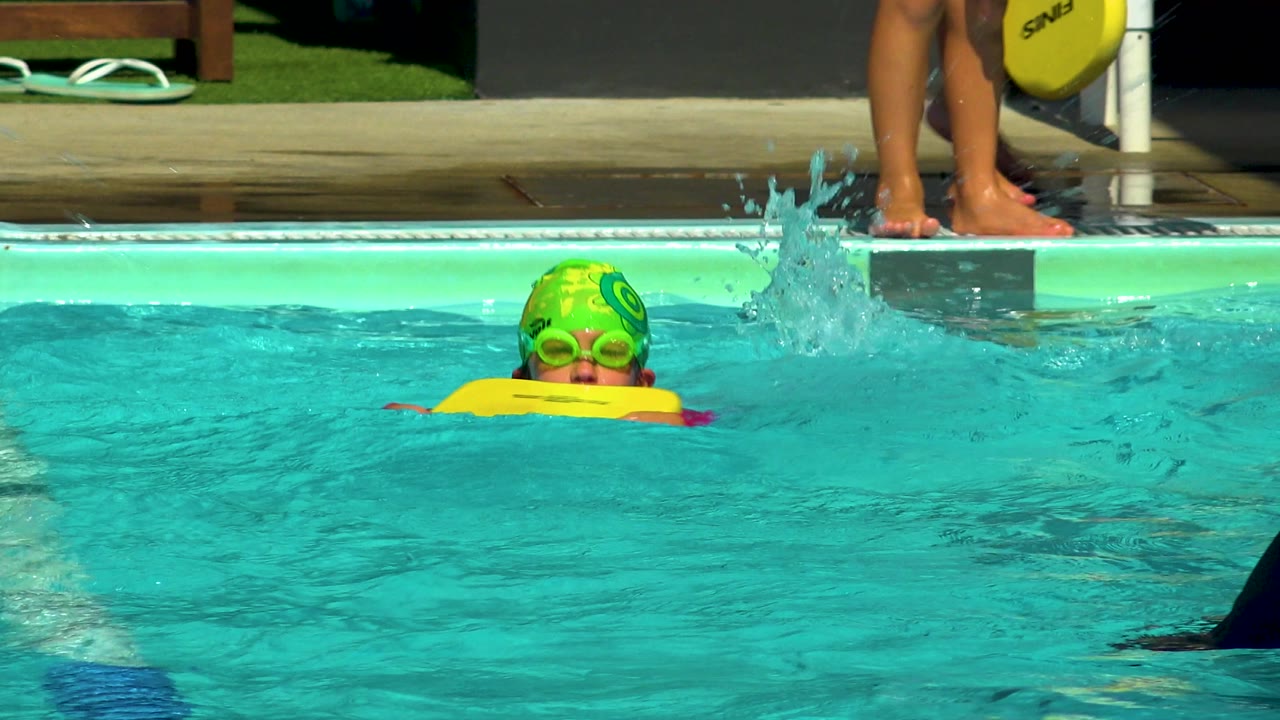 At 0:01, the child remains upright behind the yellow kickboard as splashing water rises near the pool edge and feet on the deck remain visible.