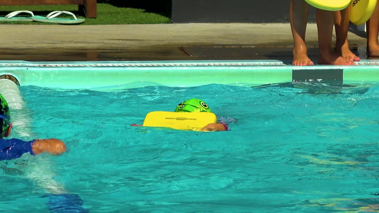 At 0:02, the child is farther across the pool with face partly down behind the yellow kickboard, a lane divider running through the water, and pool-edge legs in the background.
