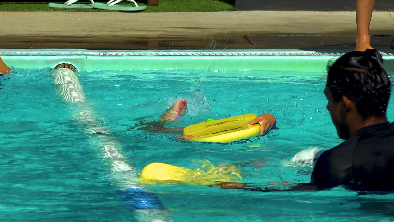 In Cabo San Lucas, a child swimmer is partly submerged while holding a yellow kickboard, with an adult instructor in a black swim shirt in the foreground.