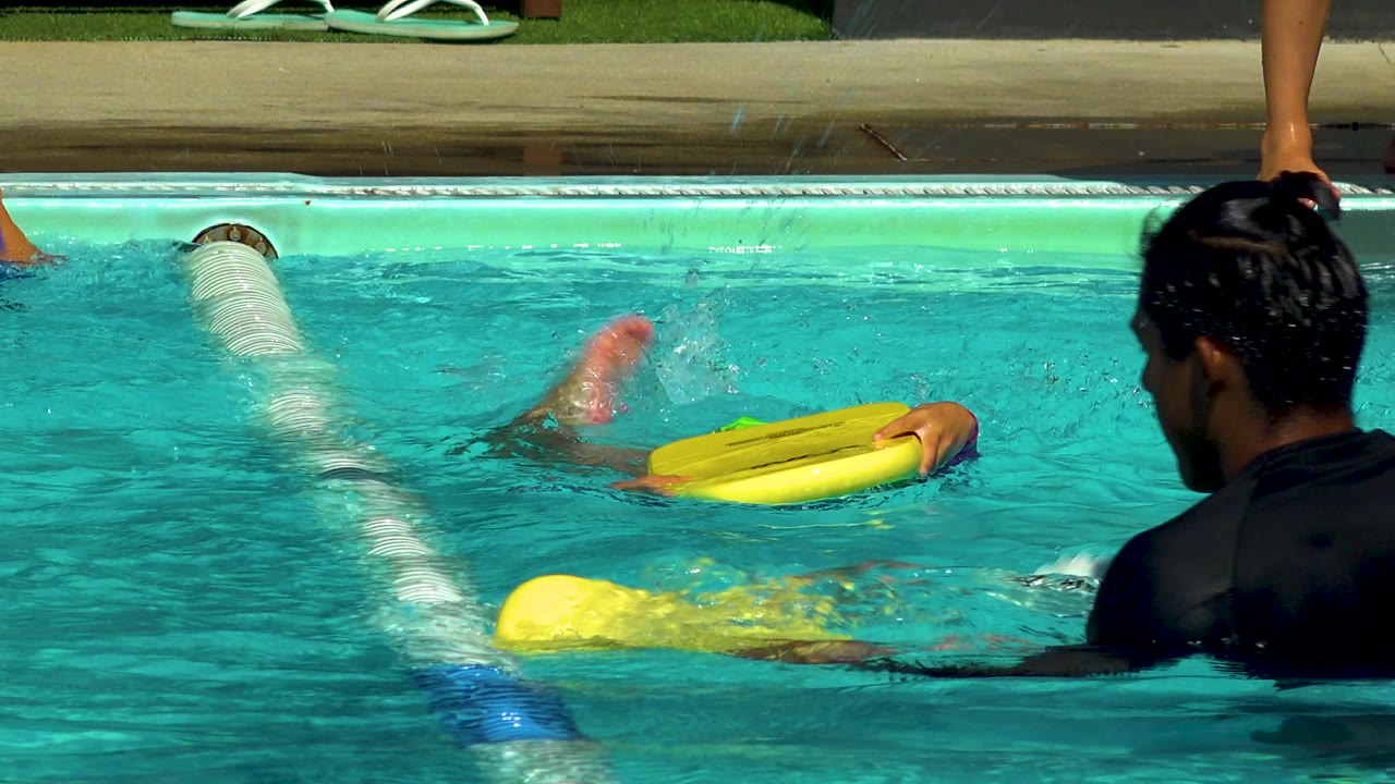 Child kicks through pool water while holding a yellow float board.