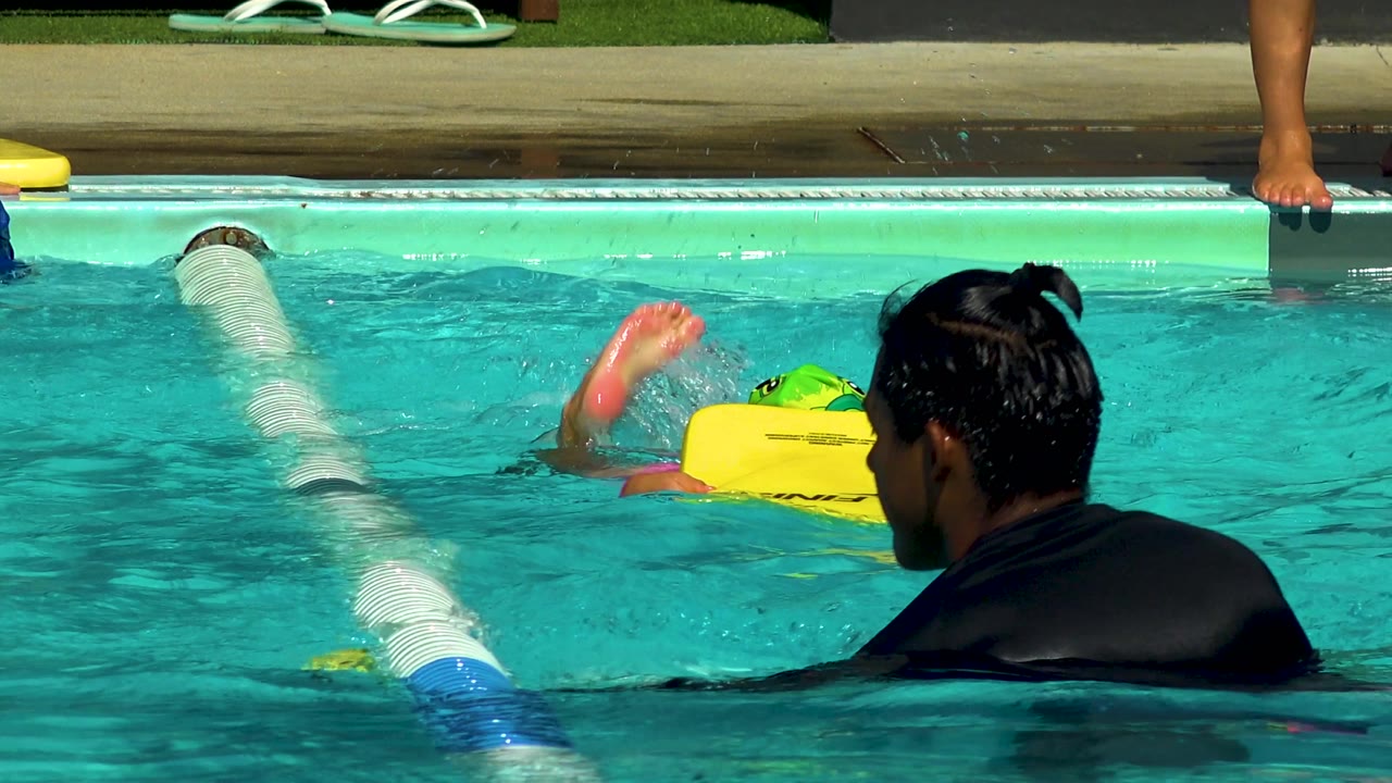 In Cabo San Lucas, a child in a green swim cap and goggles holds a yellow kickboard near a white lane divider while the adult instructor watches from the right foreground.