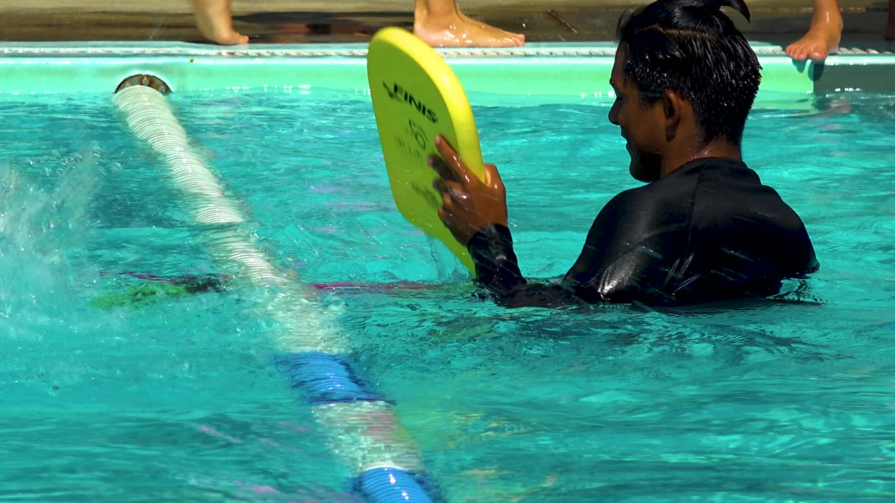 In Cabo San Lucas, Mexico, a man in a black swim shirt stands in turquoise pool water holding a yellow kickboard beside a white lane divider.