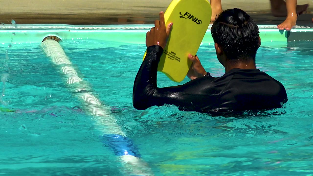 From behind, the man holds the yellow kickboard above the water while the bright pool surface and lane rope fill the frame in Cabo San Lucas, Mexico.