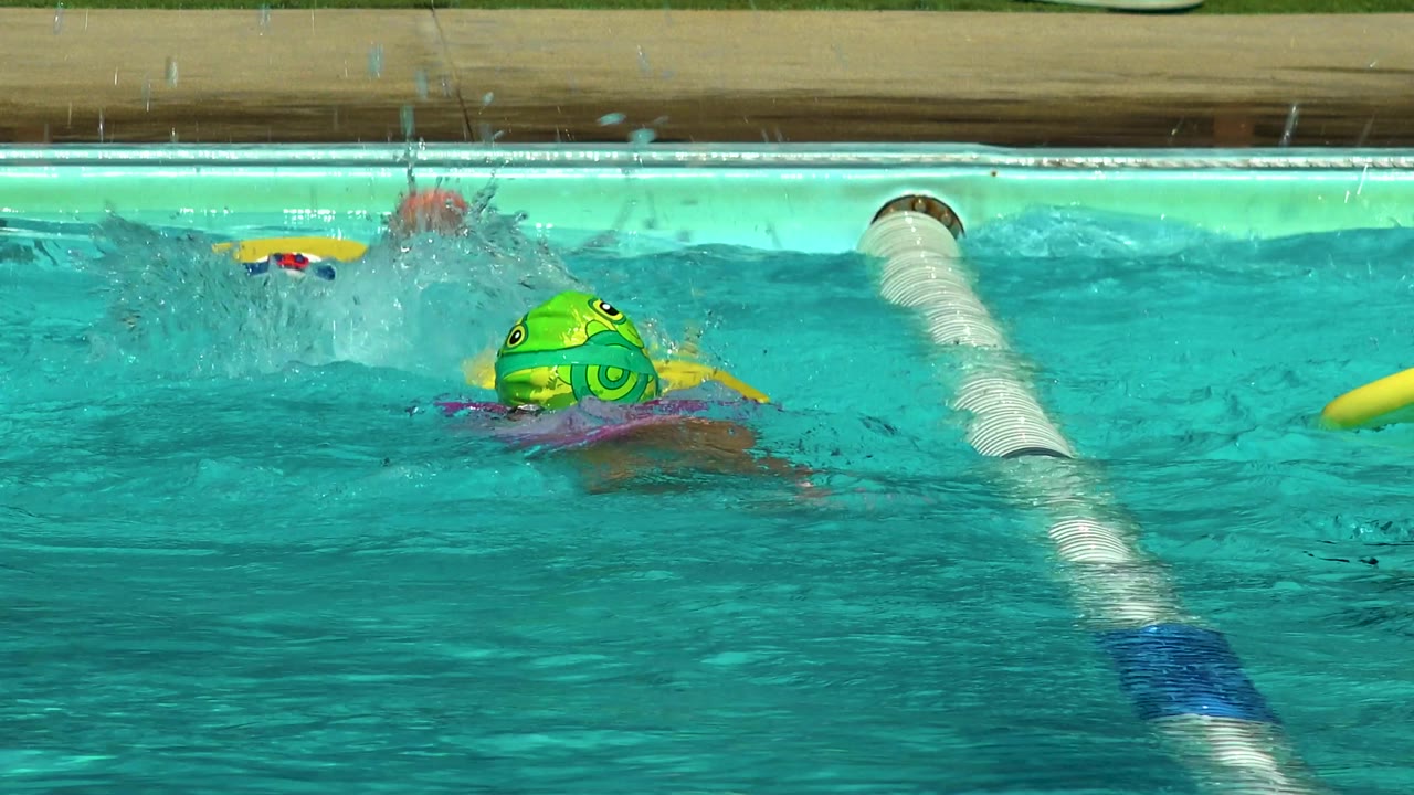 A child wearing a bright green swim cap swims near a white-and-blue lane rope in the Cabo San Lucas pool while water splashes above the lane.