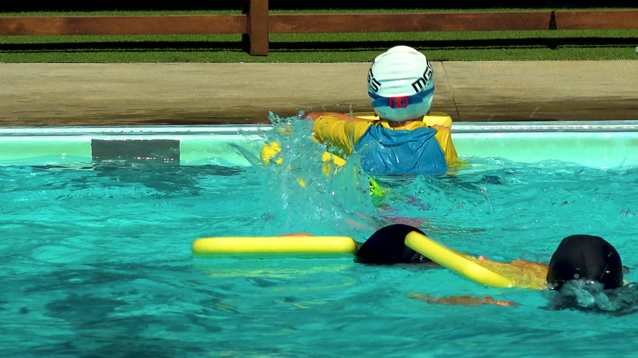 At a Cabo San Lucas pool, children in swim caps use yellow flotation boards while a child in a blue and yellow rash guard sits at the pool edge with their back to camera.