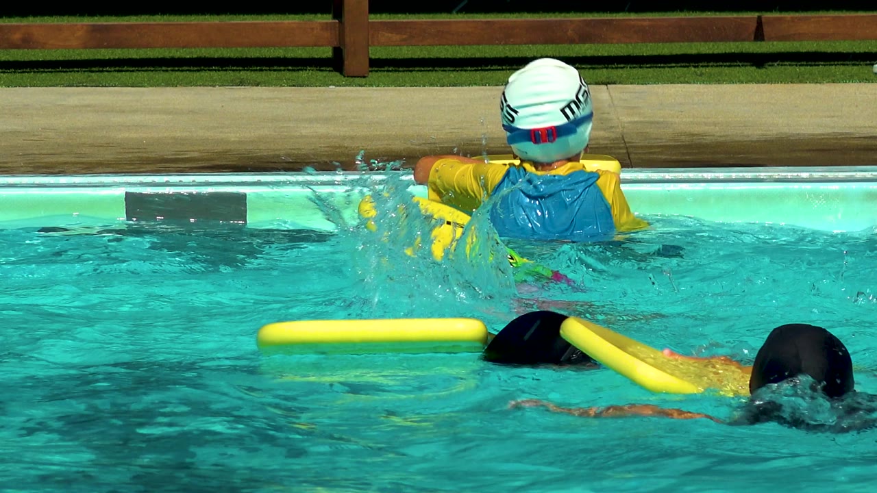 Swimmers splash near yellow foam flotation boards in a pool.