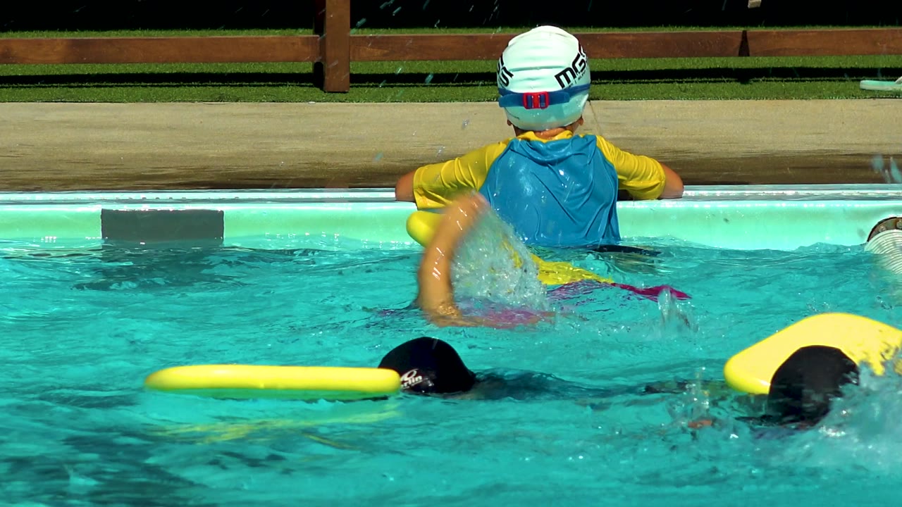 A child in a white swim cap and blue vest sits on the Cabo San Lucas pool ledge while children with yellow flotation boards swim across the foreground.
