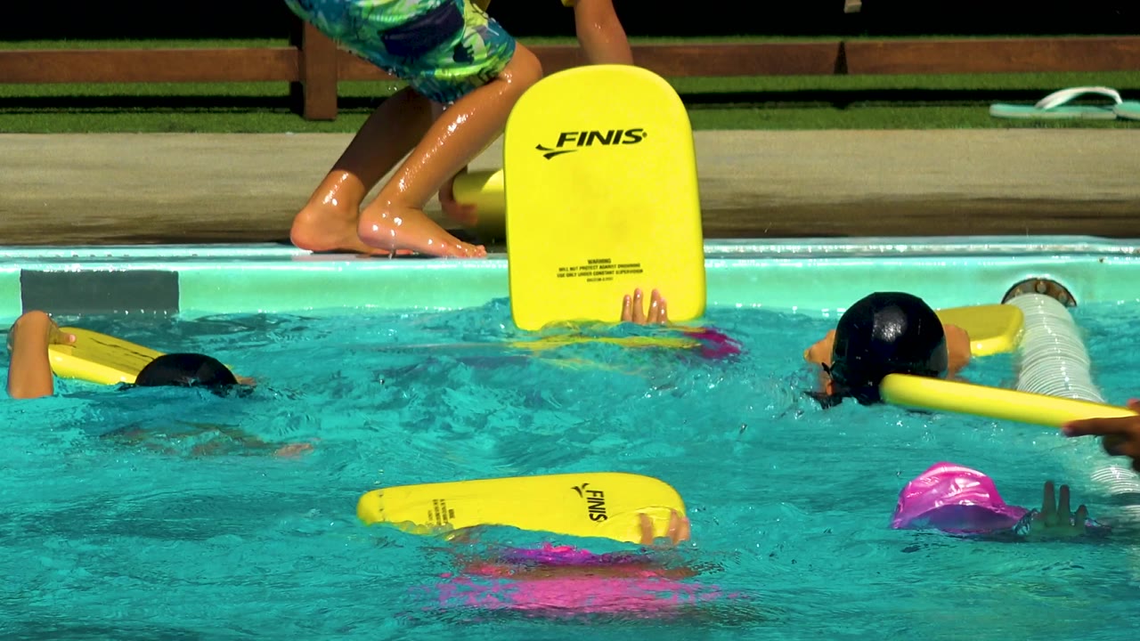 A child lifts a yellow kickboard at the right side of the Cabo San Lucas pool while other children swim with flotation aids nearby.
