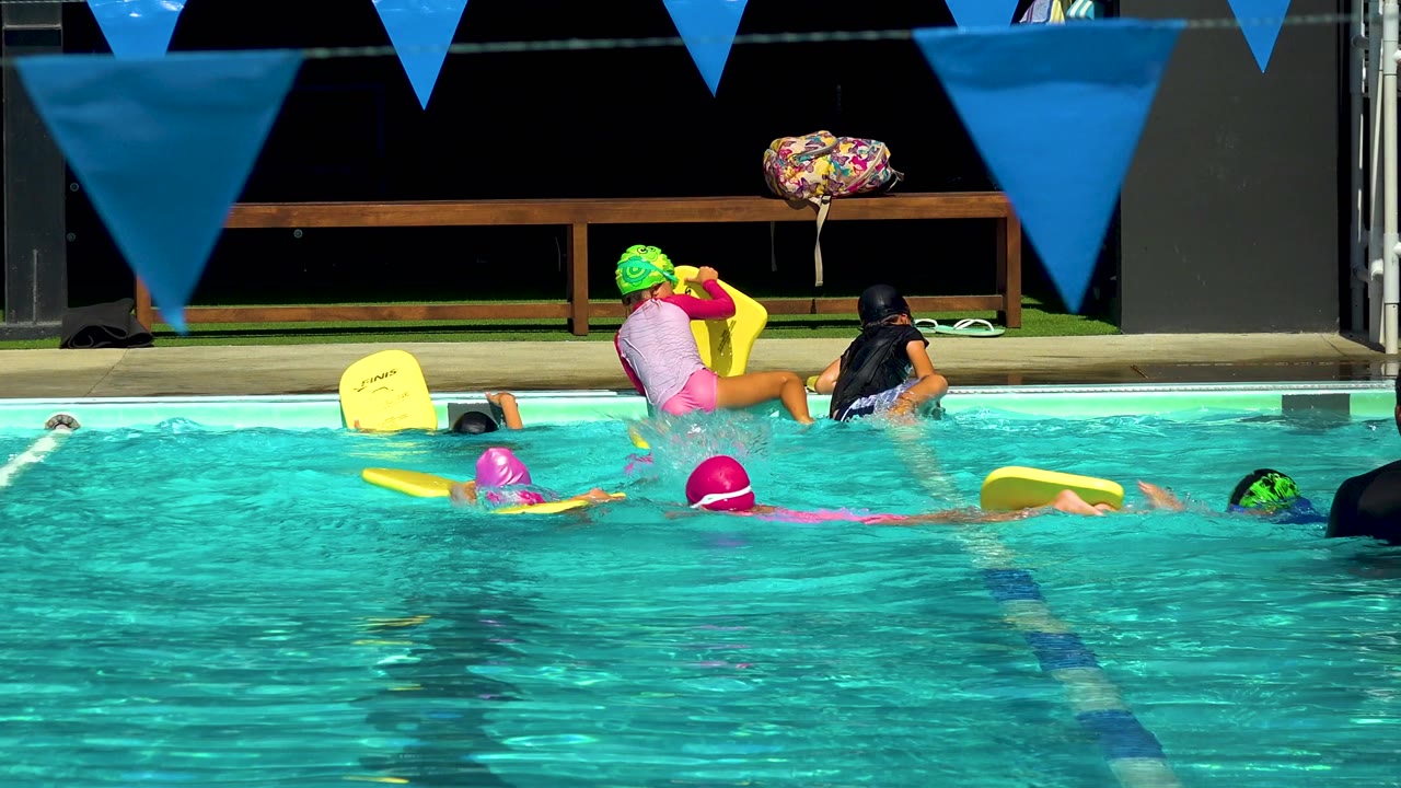 Children practice with yellow kickboards in a turquoise pool in Cabo San Lucas, Mexico, with blue pennants overhead and an instructor near the far deck.