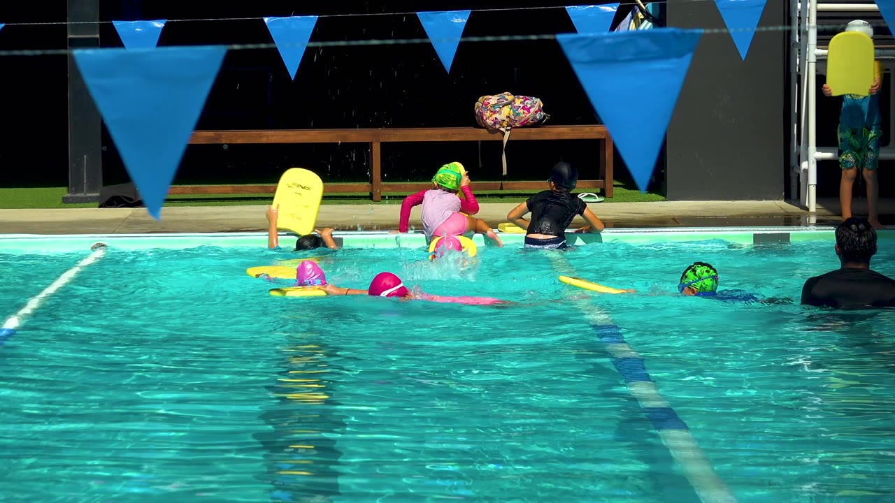 Children and instructors cluster closer to the pool edge in Cabo San Lucas, Mexico, as yellow kickboards float near the lane lines under blue triangular pennants.