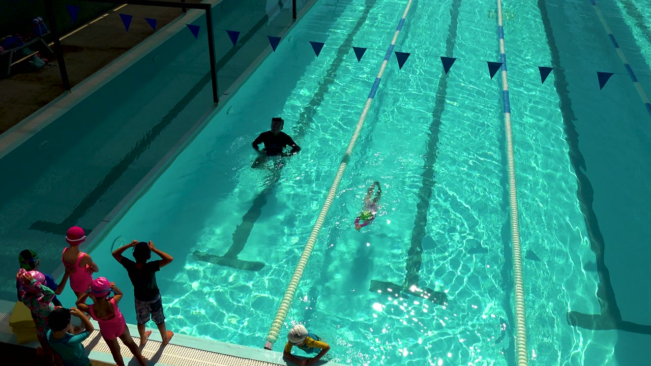 In Cabo San Lucas, Mexico, a child swims through a turquoise pool lane while an adult stands farther ahead in the water and a small group waits at the deck edge.