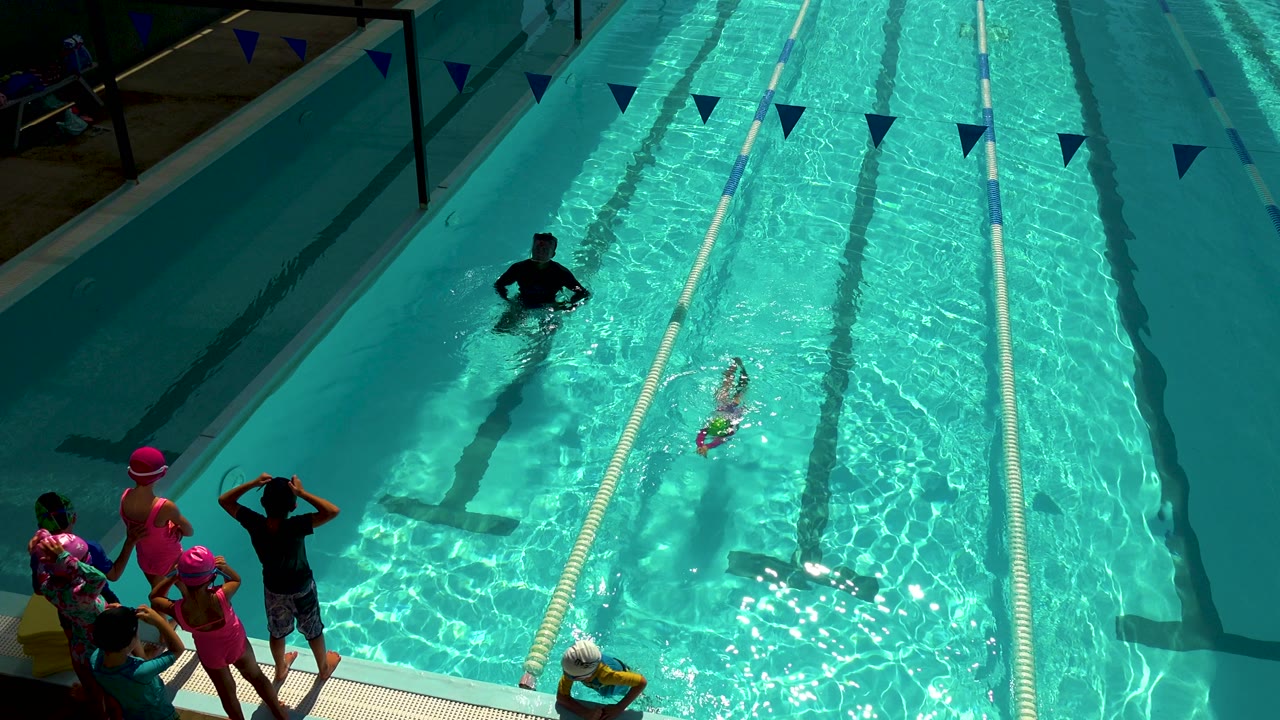 Child swimmer moves between lane ropes with a group gathered at poolside.