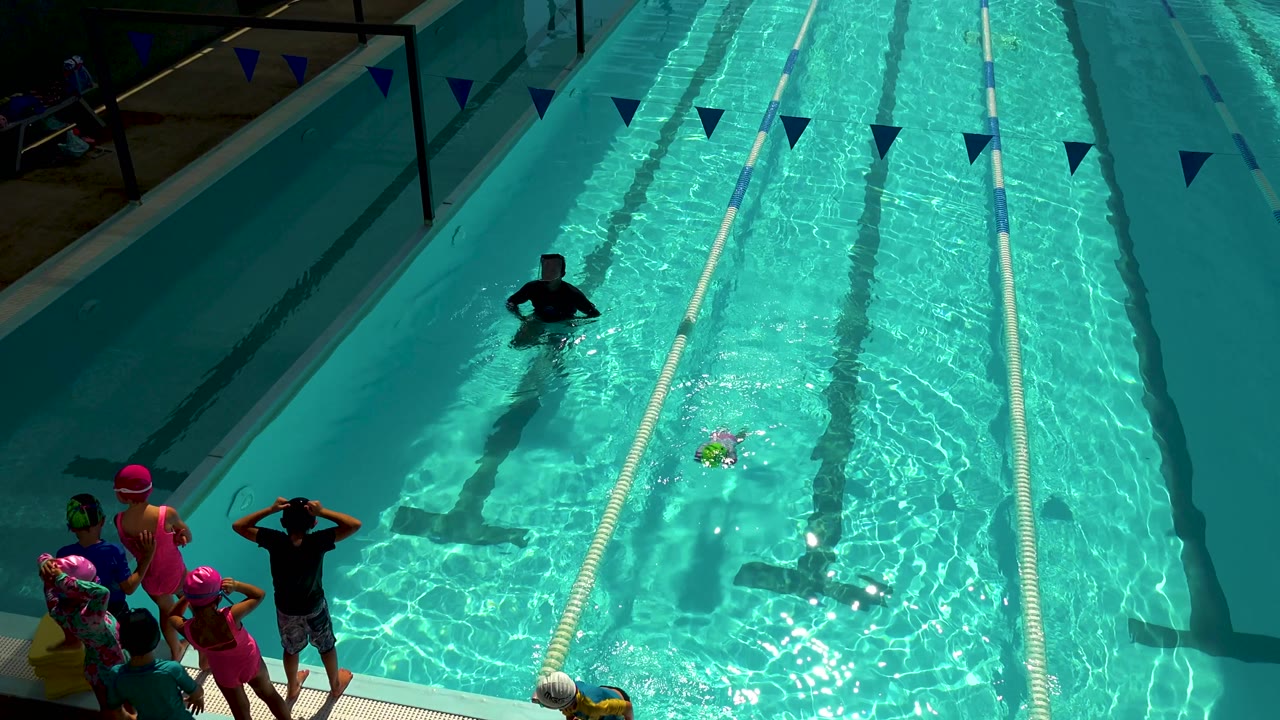 In Cabo San Lucas, Mexico, the child in a bright cap and pink sleeves is farther down the lane while the adult in dark clothing stands in the pool beyond the swimmer.