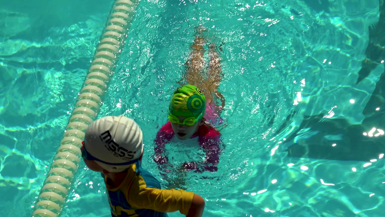 In Cabo San Lucas, a young girl in a green swim cap swims toward the camera while another child in a white cap is partly visible beside the lane rope.