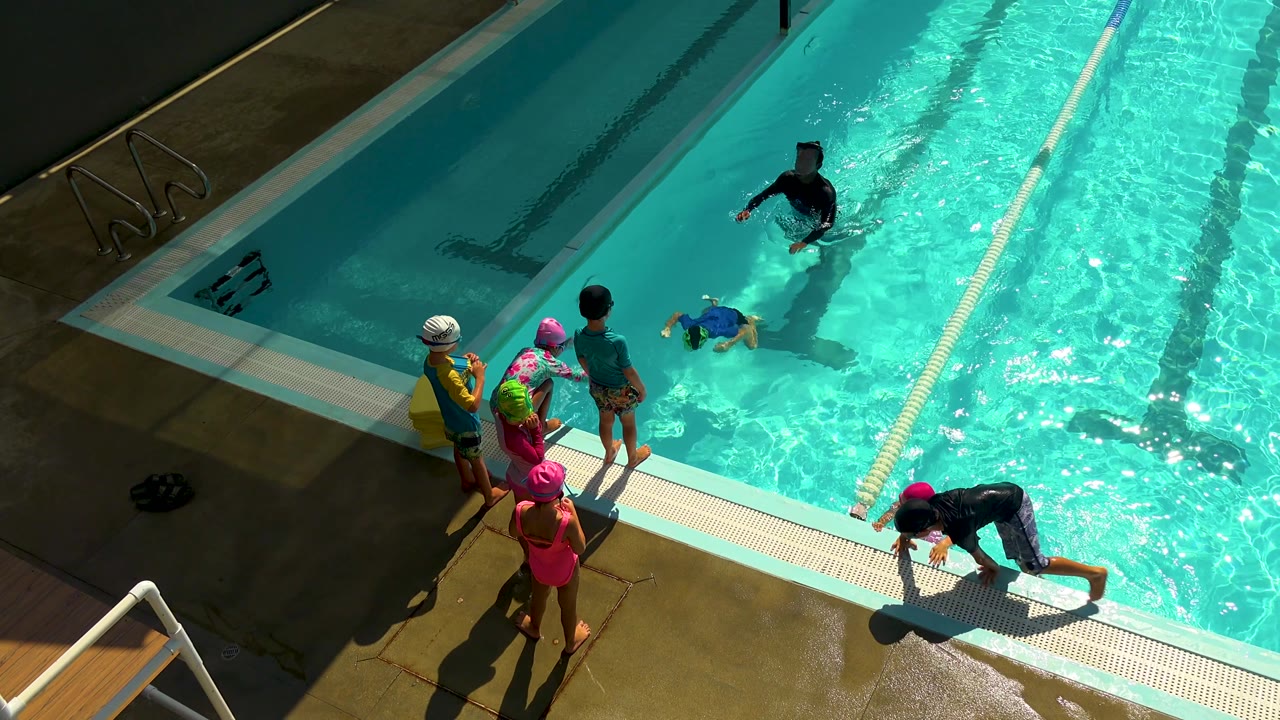 At 0:02 in Cabo San Lucas, Mexico, one child climbs onto the pool edge as the instructor and the waiting children hold position around the bright blue water.