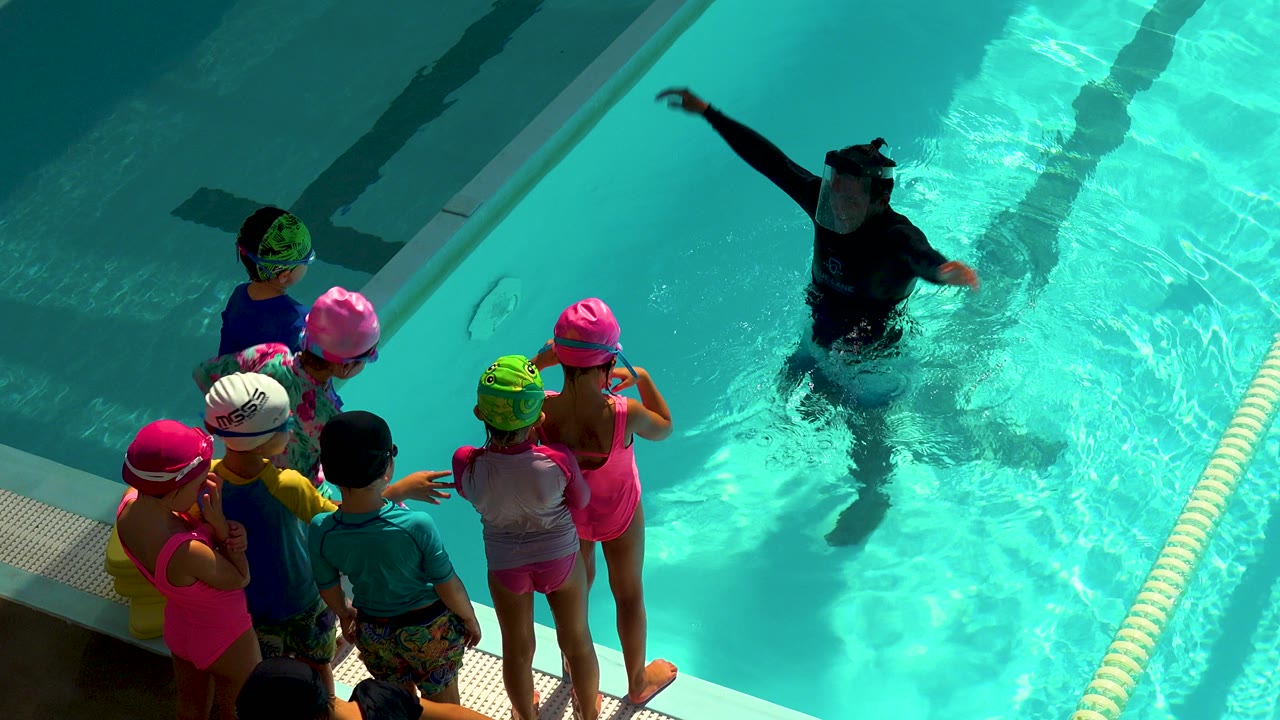 Children in bright swim caps stand at a pool edge in Cabo San Lucas, Mexico while an adult instructor demonstrates with one arm raised in the water.