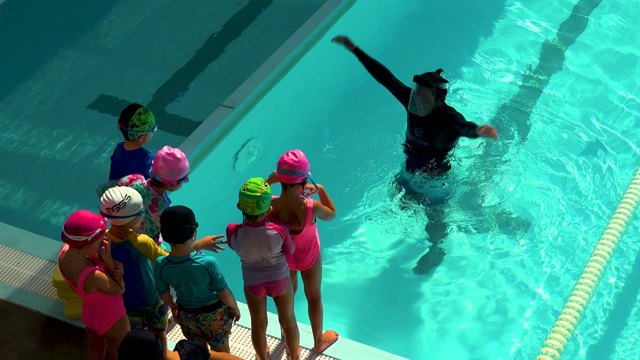 Children stand beside a pool while an instructor gestures in the water.