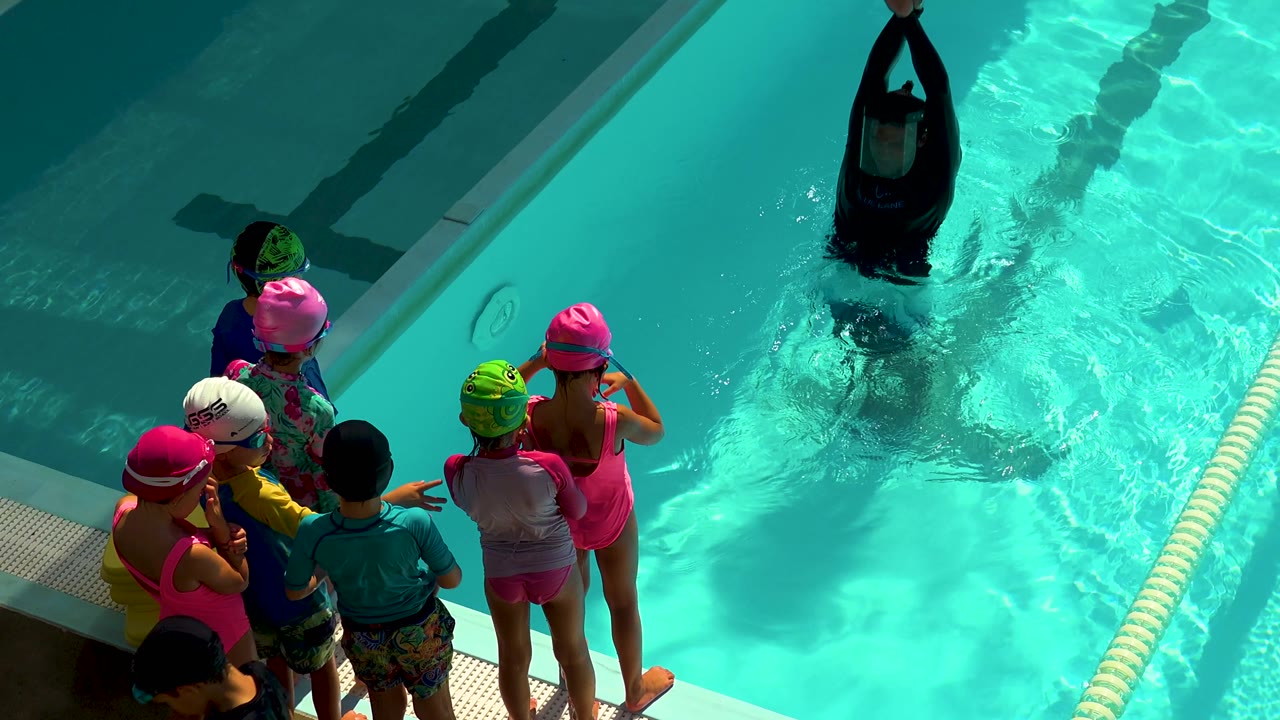 The instructor lifts both arms above the water while children in swim caps line the pool edge in Cabo San Lucas, Mexico.