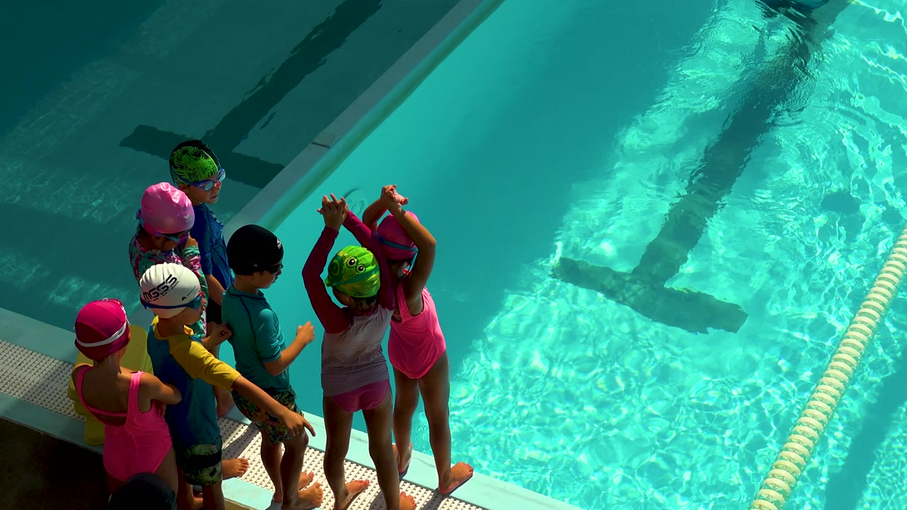 Children in colorful swim caps and swimsuits stand on the pool deck in Cabo San Lucas, Mexico, looking toward the bright turquoise water.
