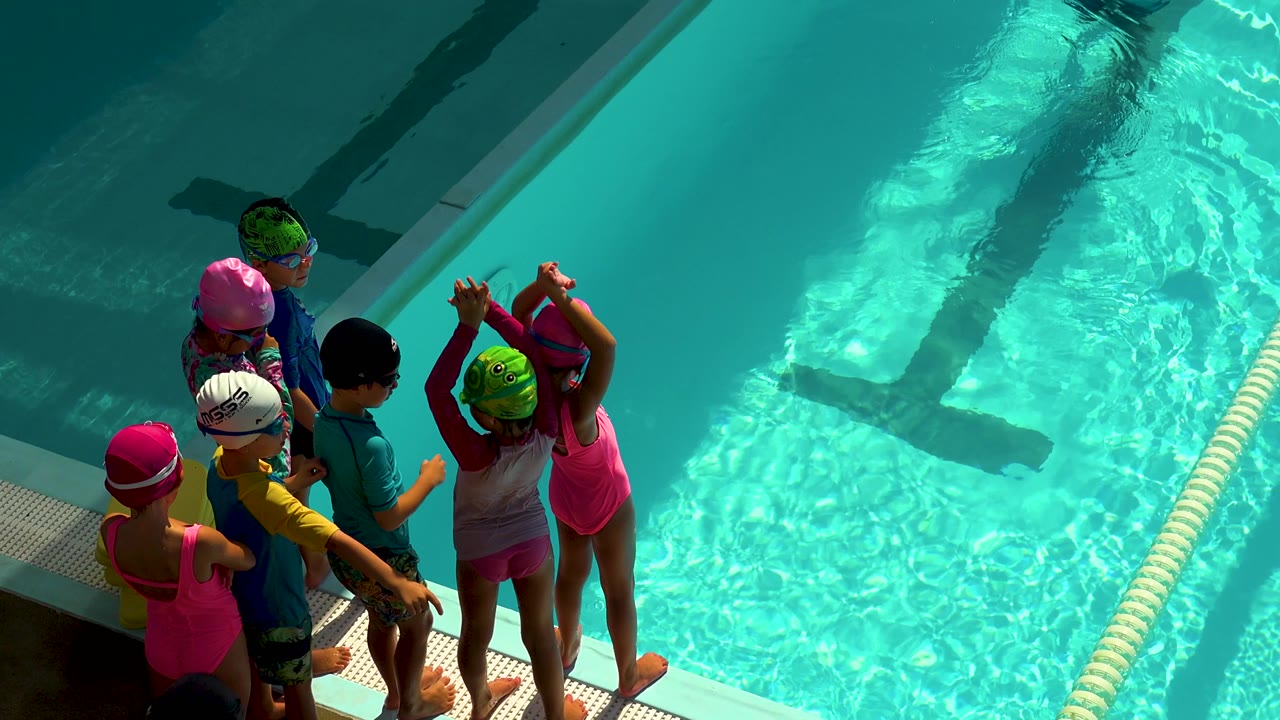 Children wait together beside a bright swimming pool.