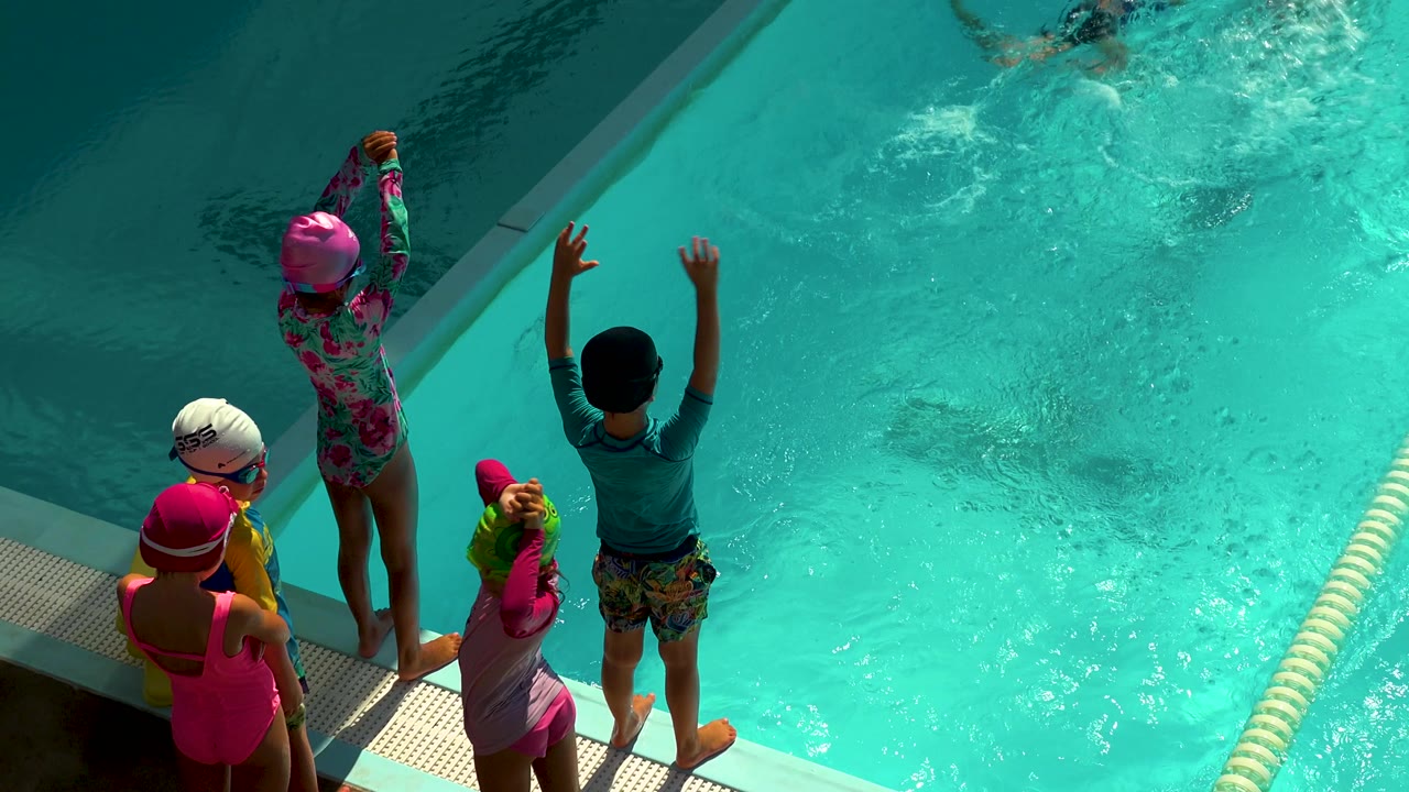 Children in colorful swim caps stand along a pool edge in Cabo San Lucas, Mexico, with one child raising an arm toward the turquoise water.