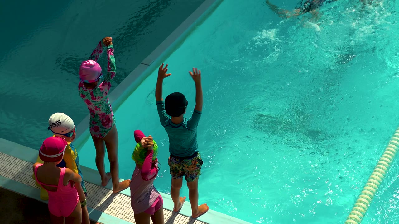Children prepare at the edge of a bright blue swimming pool.