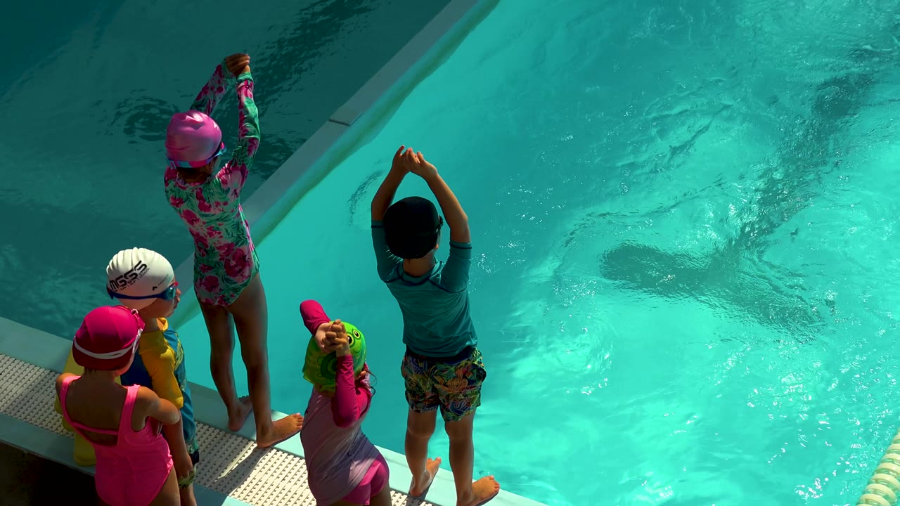 Children at the pool edge in Cabo San Lucas, Mexico watch the water, with one child in blue holding both arms above the head and another in a floral suit raising an arm.