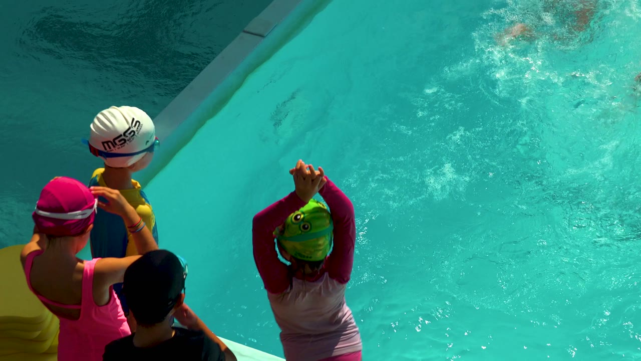 Children in swim caps and goggles stand at the edge of a turquoise pool in Cabo San Lucas, Mexico, with sunlit water filling the frame.