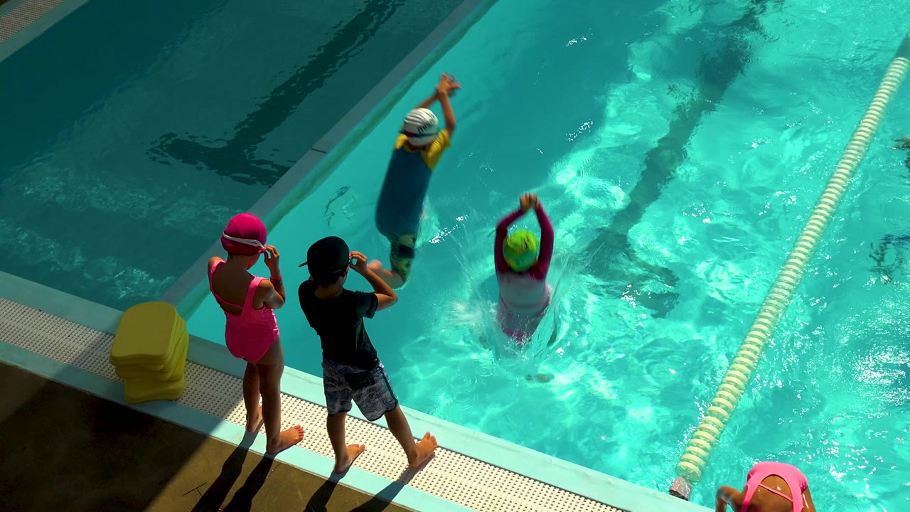 High-angle view in Cabo San Lucas, Mexico, showing children at the pool edge as two swimmers move through turquoise water with kickboards and a lane rope beside them.