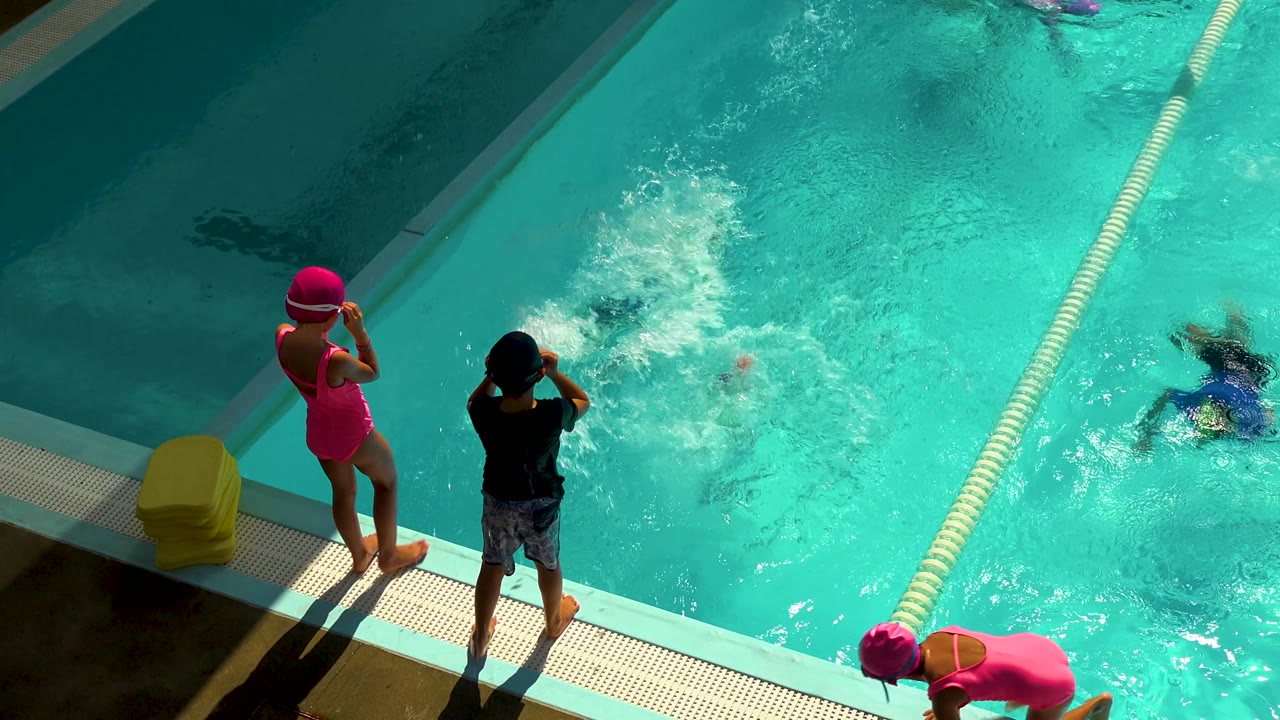 Two children remain on the deck in Cabo San Lucas, Mexico, while splashing water, swimmers, a lane rope, and a child leaning near the lower edge show active pool play.