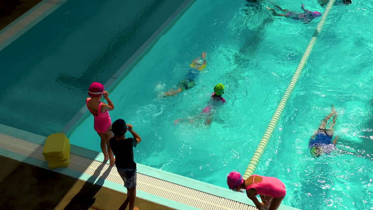 Children swim in a bright pool while two children stand near the edge.