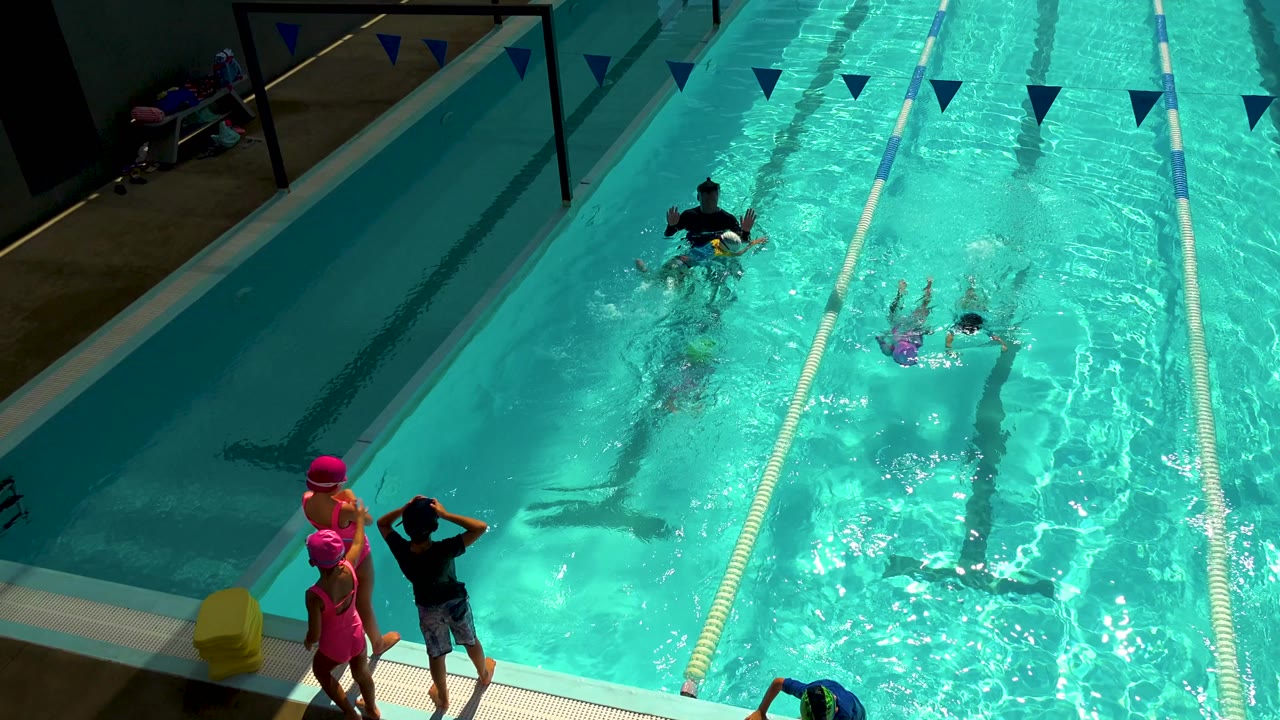 High-angle view in Cabo San Lucas, Mexico, showing children in pink caps at the pool edge, a coach in the water, and young swimmers moving between lane ropes.