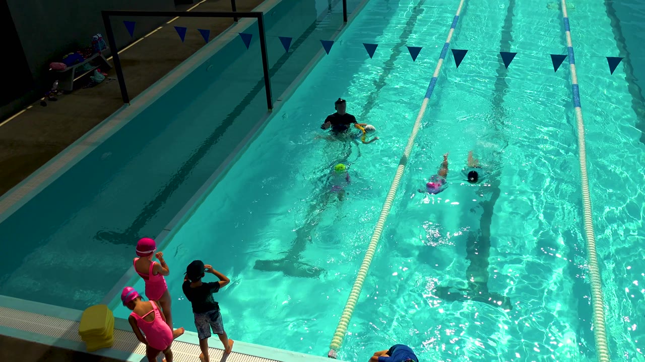 Overhead pool view in Cabo San Lucas shows two children swimming forward, a coach watching nearby, and deck-side children in pink swim caps at the lower left.
