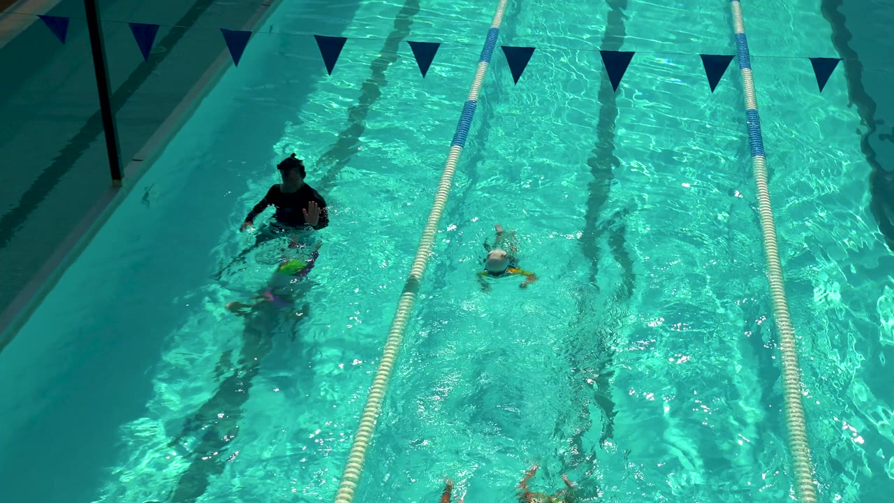Overhead view in Cabo San Lucas of a man and children in a turquoise lane pool, with pennant flags across the top and sun ripples on the water.