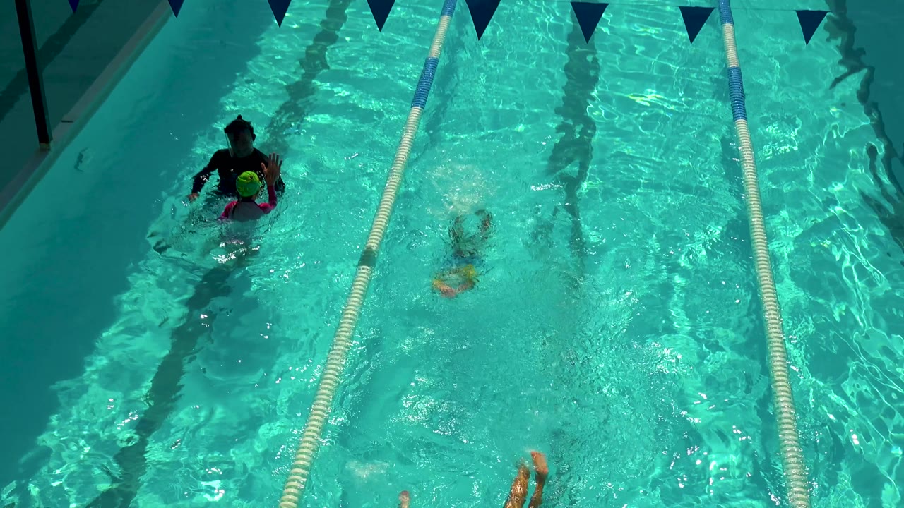 In the Cabo San Lucas pool, the adult stays near the upper left while a child with bright green and pink swim gear splashes in the neighboring lane.