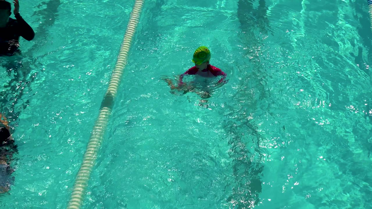 A child in a green swim cap and pink top is visible in turquoise pool water between lane ropes in Cabo San Lucas, Mexico.