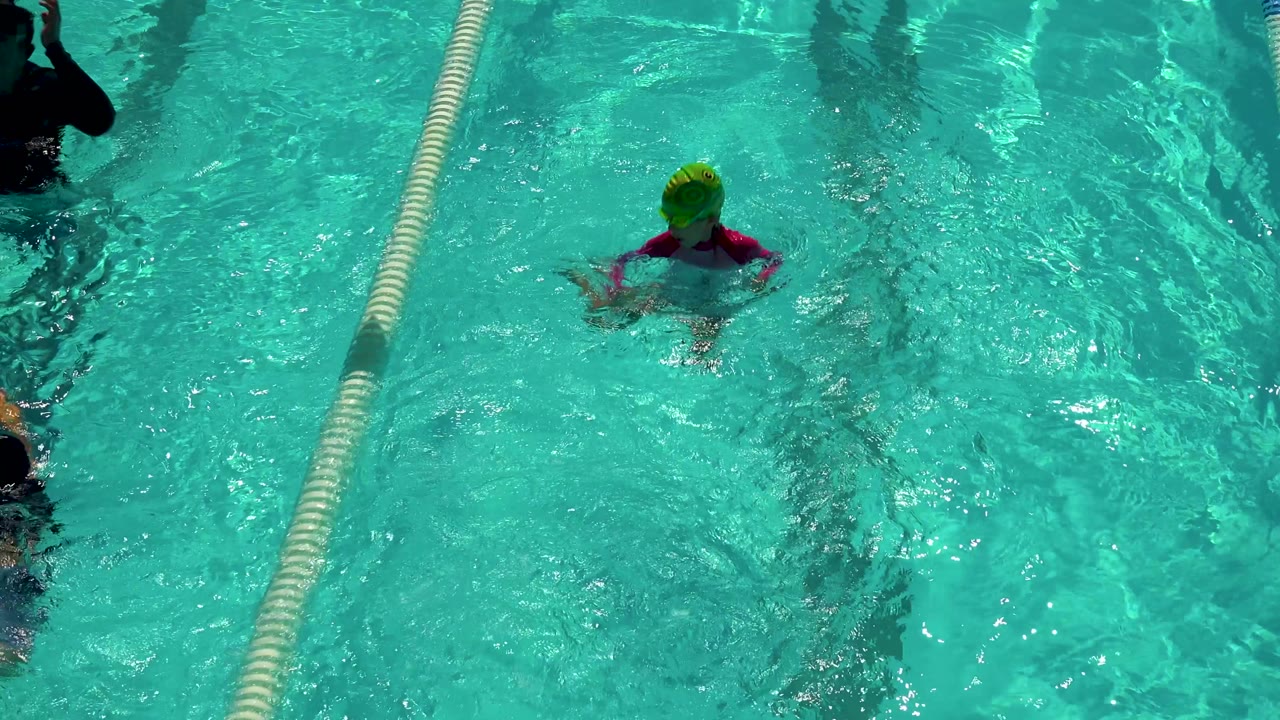 Child in a green swim cap near a lane rope in bright pool water.