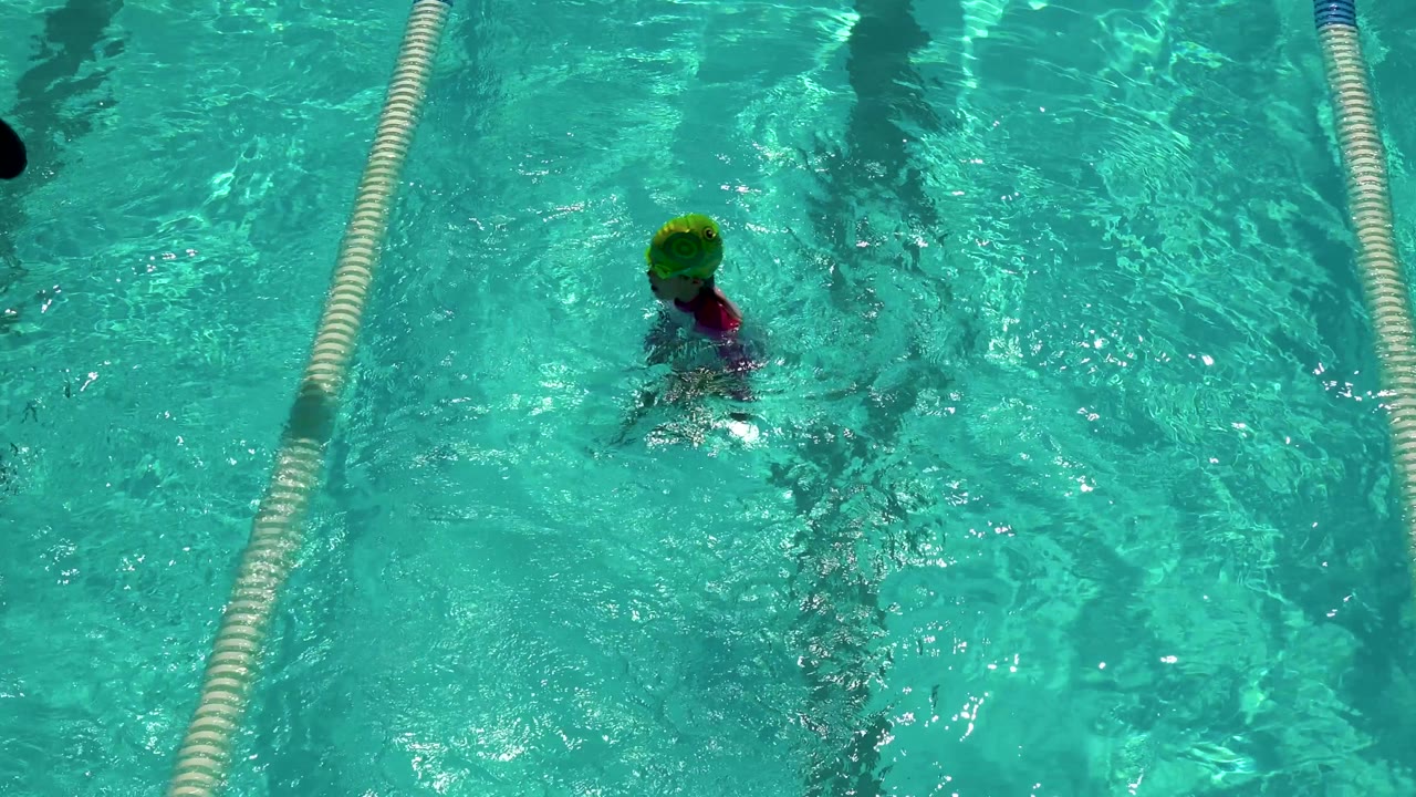 A small child in a green swim cap appears higher in the lane as the water brightens with sun reflections and lane ropes frame the swimming area in Cabo San Lucas, Mexico.