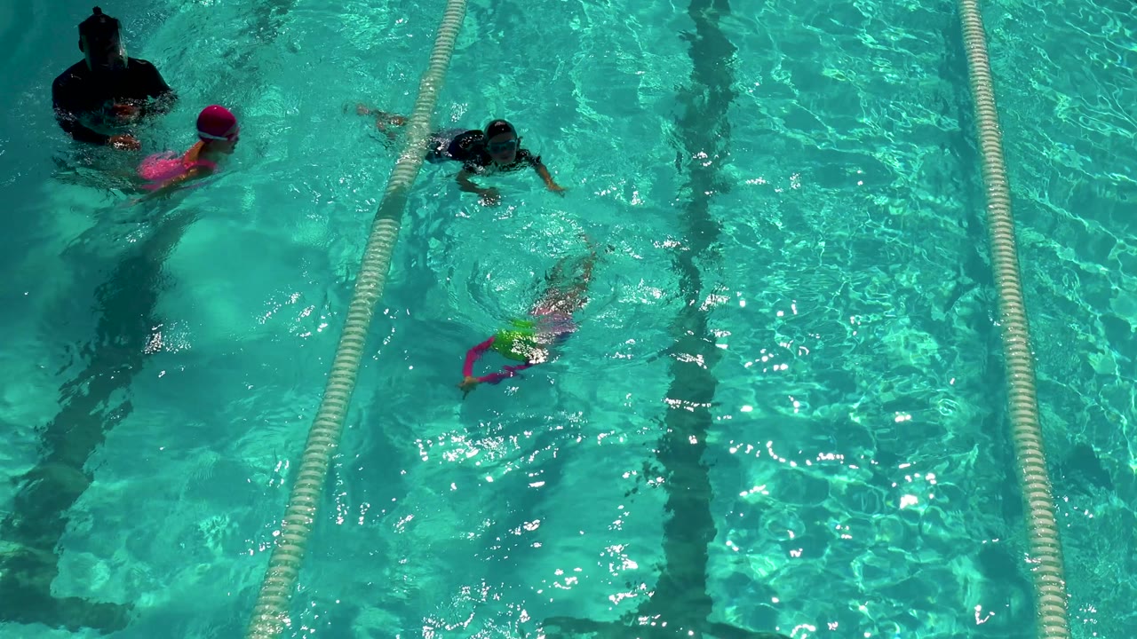 Overhead view in Cabo San Lucas, Mexico, of children in bright swim caps moving through turquoise pool lanes while an adult in dark swimwear stands at the upper left.