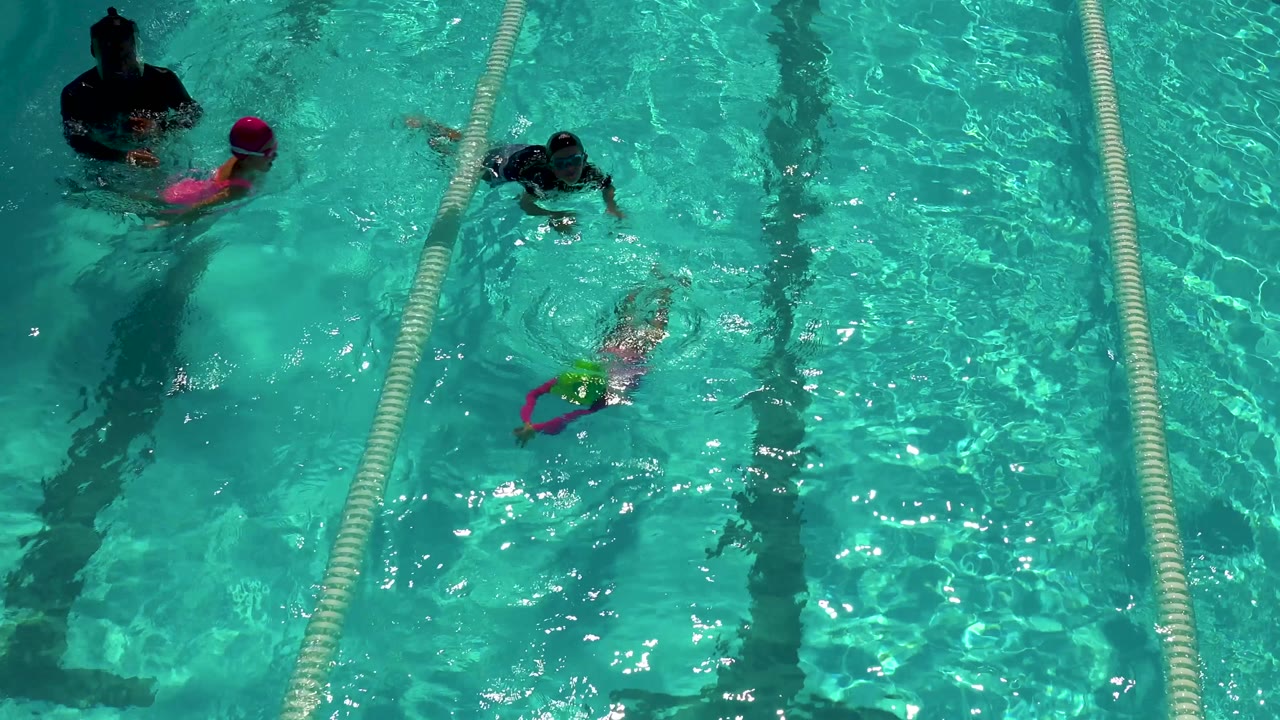 Overhead Cabo San Lucas pool frame with two children moving through adjacent lanes, one in a pink cap and another in darker swim gear, as sunlight ripples across the water.