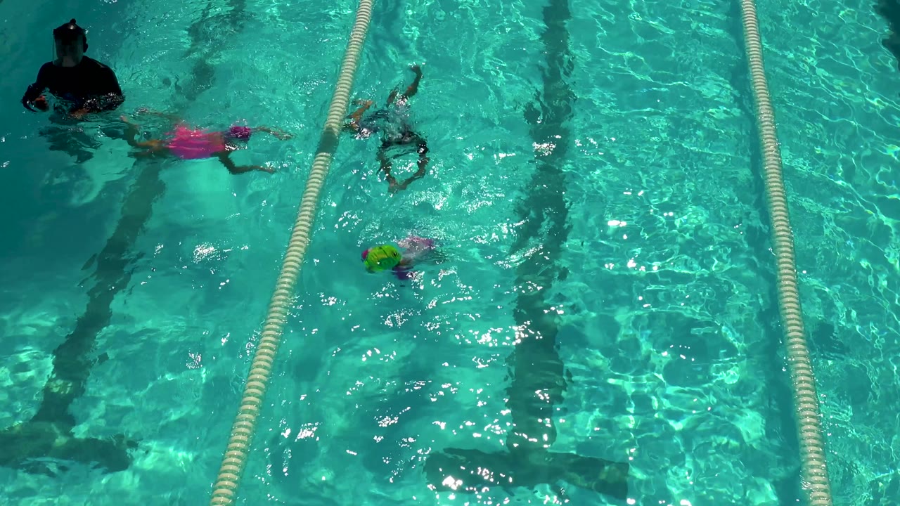 High-angle frame in Cabo San Lucas, Mexico, showing children separated across pool lanes, a pink-capped child near the upper lane, and an adult standing in the water at the top left.