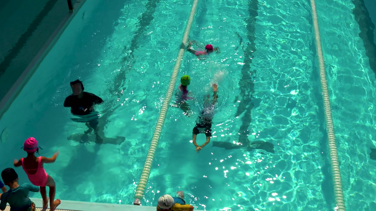 Overhead view in Cabo San Lucas of children in swim caps and an adult instructor standing in a turquoise pool beside lane ropes.