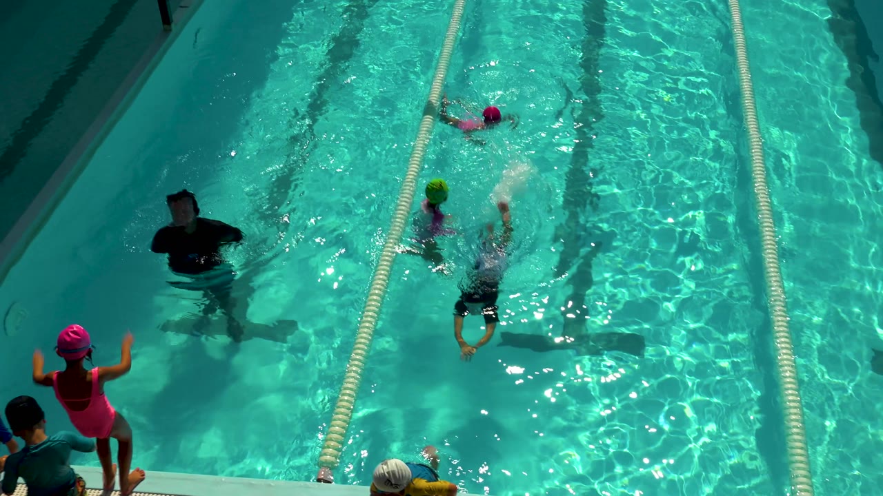Swimmers move through pool lanes while an adult stands nearby in the water.
