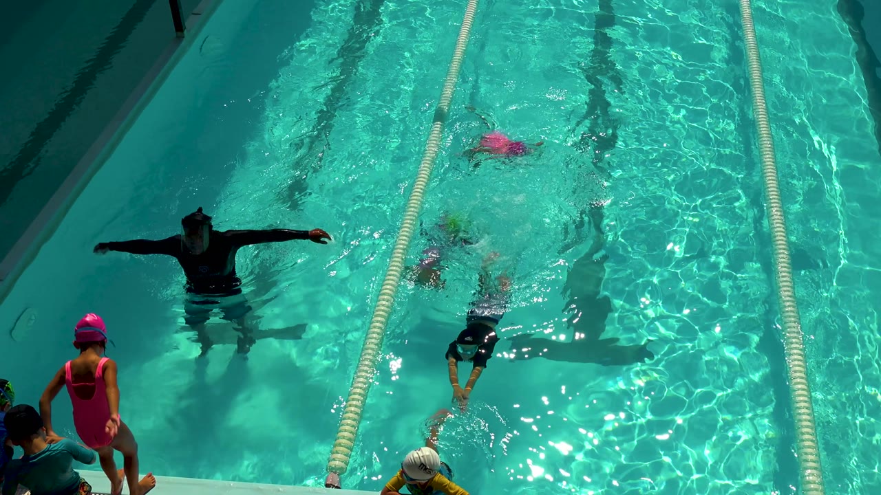 Overhead Cabo San Lucas pool frame with the adult instructor extending both arms as children swim and wait near the lane ropes.