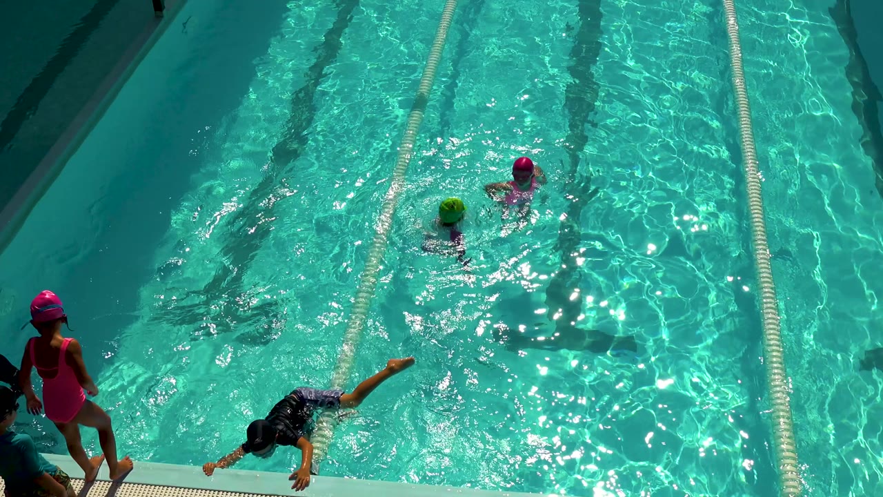 Overhead view in Cabo San Lucas, Mexico, showing girls in pink and green swim caps swimming near lane ropes while another person stands at the pool edge.