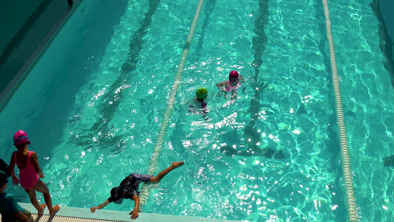 Swimmers move through sunlit pool lanes as a girl stands at the edge.