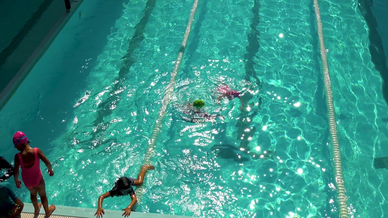 High angle in Cabo San Lucas, Mexico, with two girls swimming in adjacent pool lanes and a dark-clothed person partly visible near the lower edge.