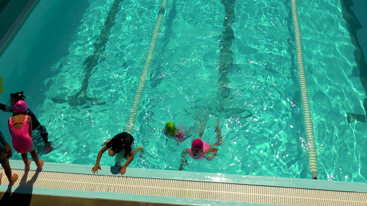 Overhead view in Cabo San Lucas, Mexico, with children in bright swim caps near the pool edge, adults on the deck, a lane divider, and sunlit turquoise water.